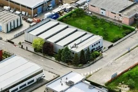 Veduta aerea di un complesso di edifici industriali con tetti bianchi, strade asfaltate e prato verde circostante.