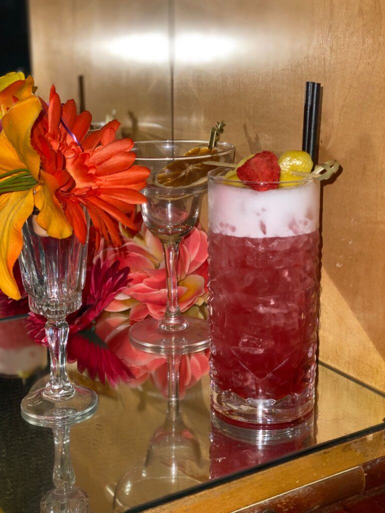 A tall glass of red liquid is sitting on a glass table next to a vase of flowers.