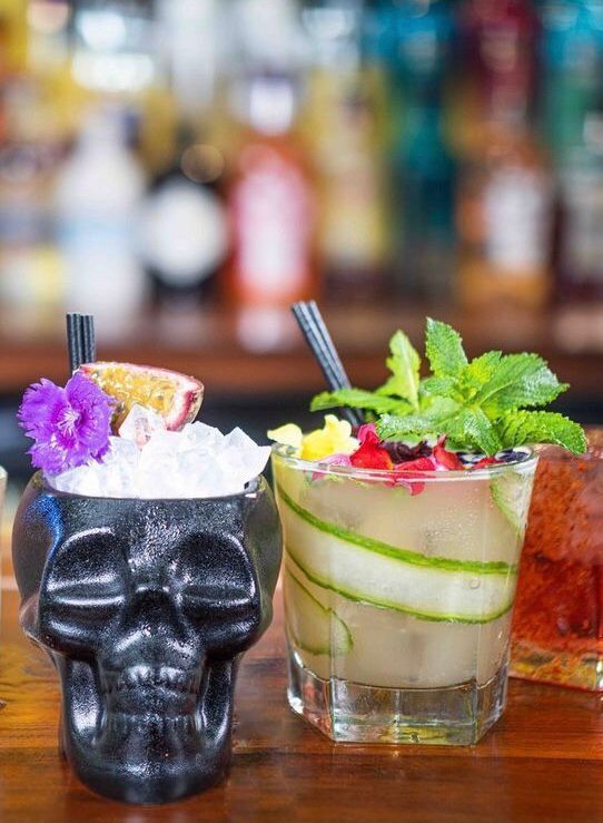 Two cocktails are sitting on a wooden table next to a skull cup.