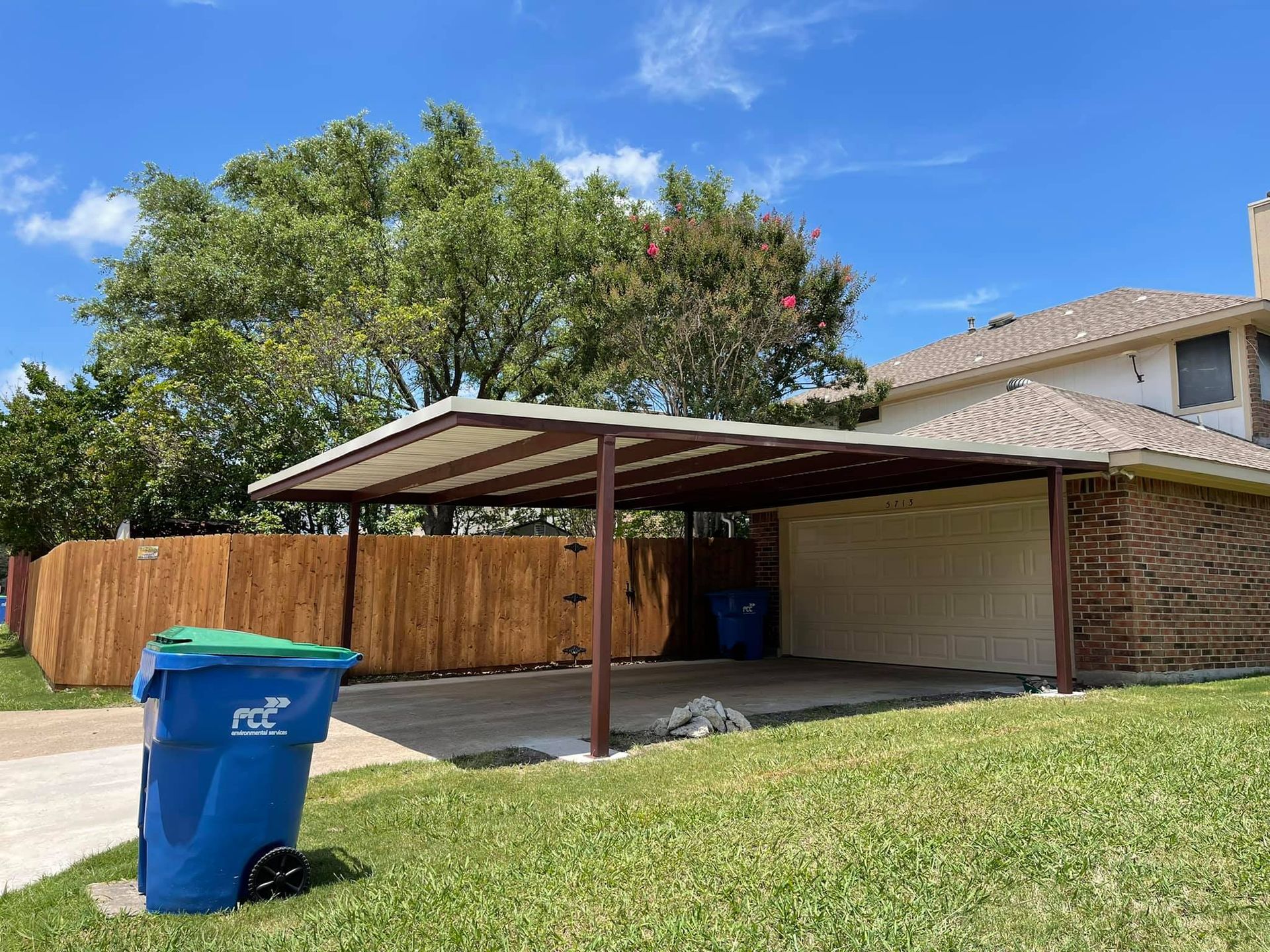A blue trash can is sitting in front of a house with a carport.
