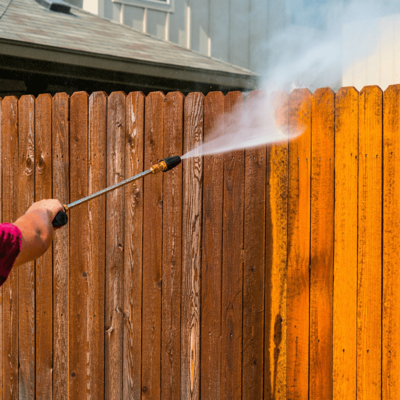 A person is cleaning a wooden fence with a high pressure washer.