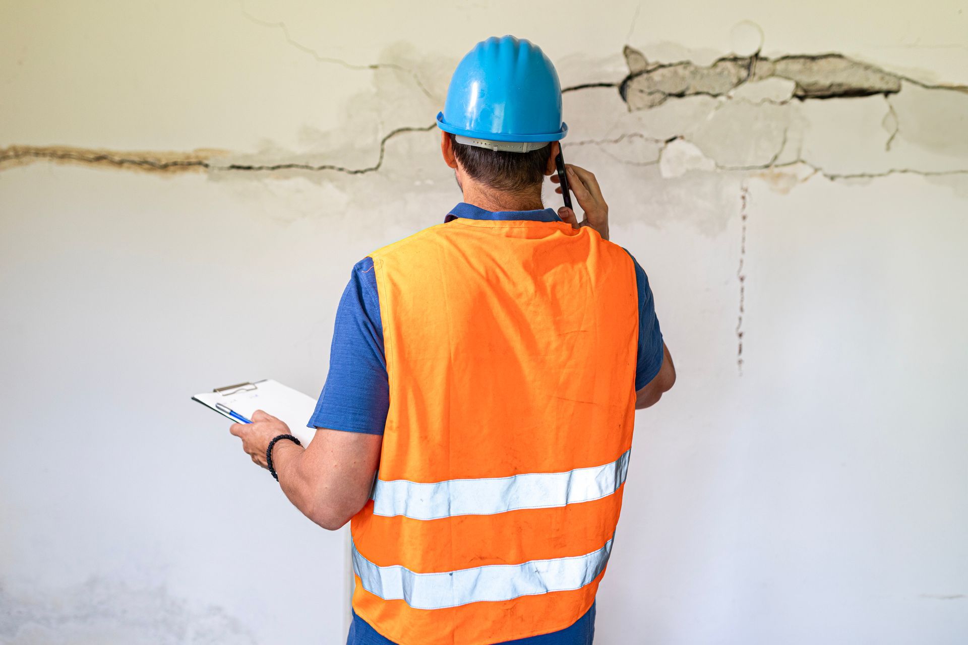A construction worker is talking on a cell phone while looking at a cracked wall.