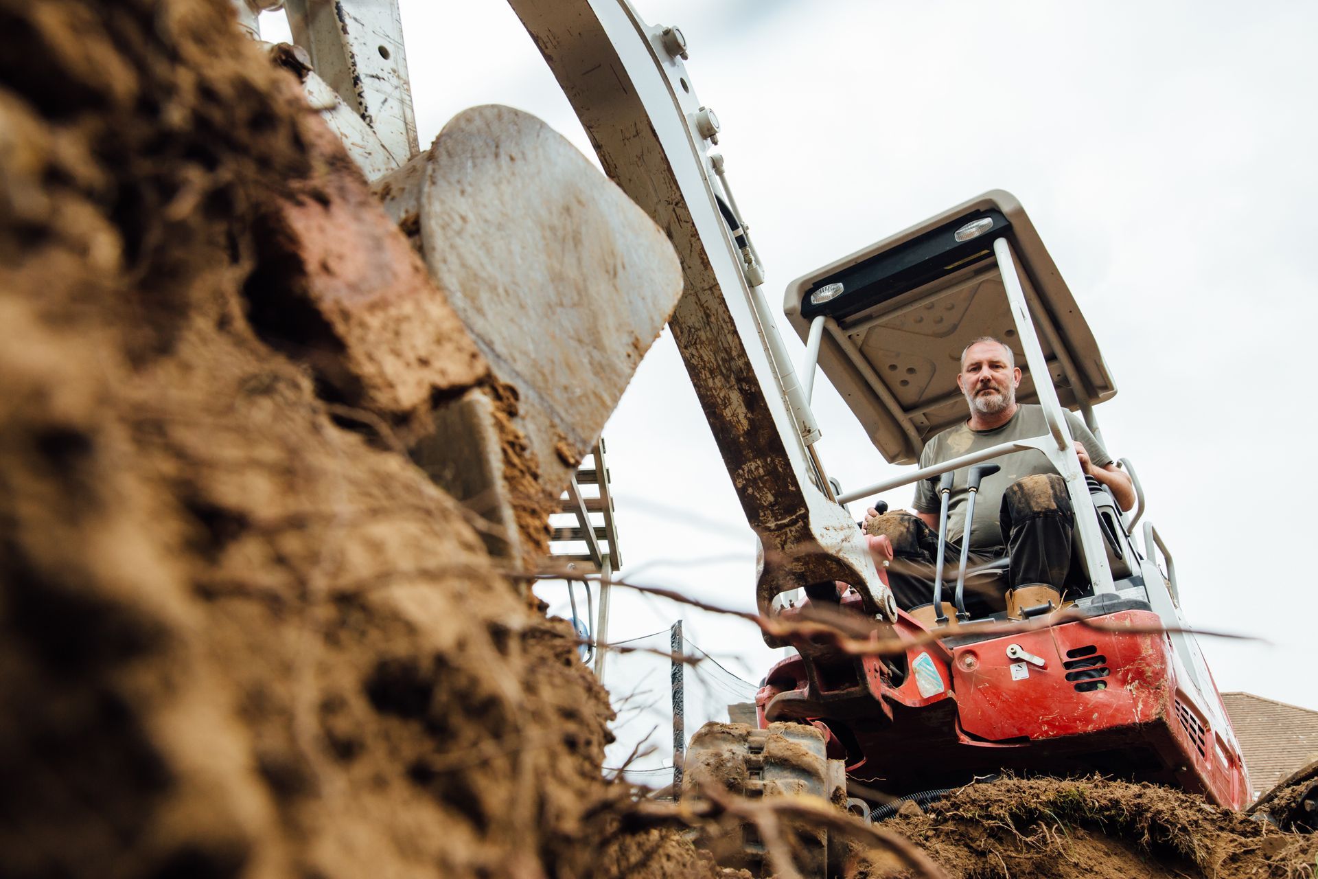 Man operating a red excavator, digging in brown dirt.