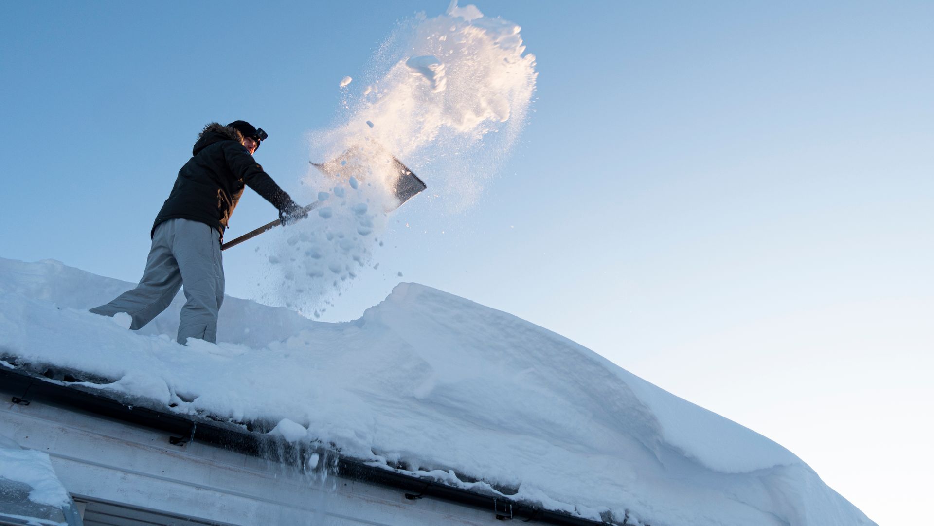 Person shoveling snow off a roof with a clear blue sky in the background.