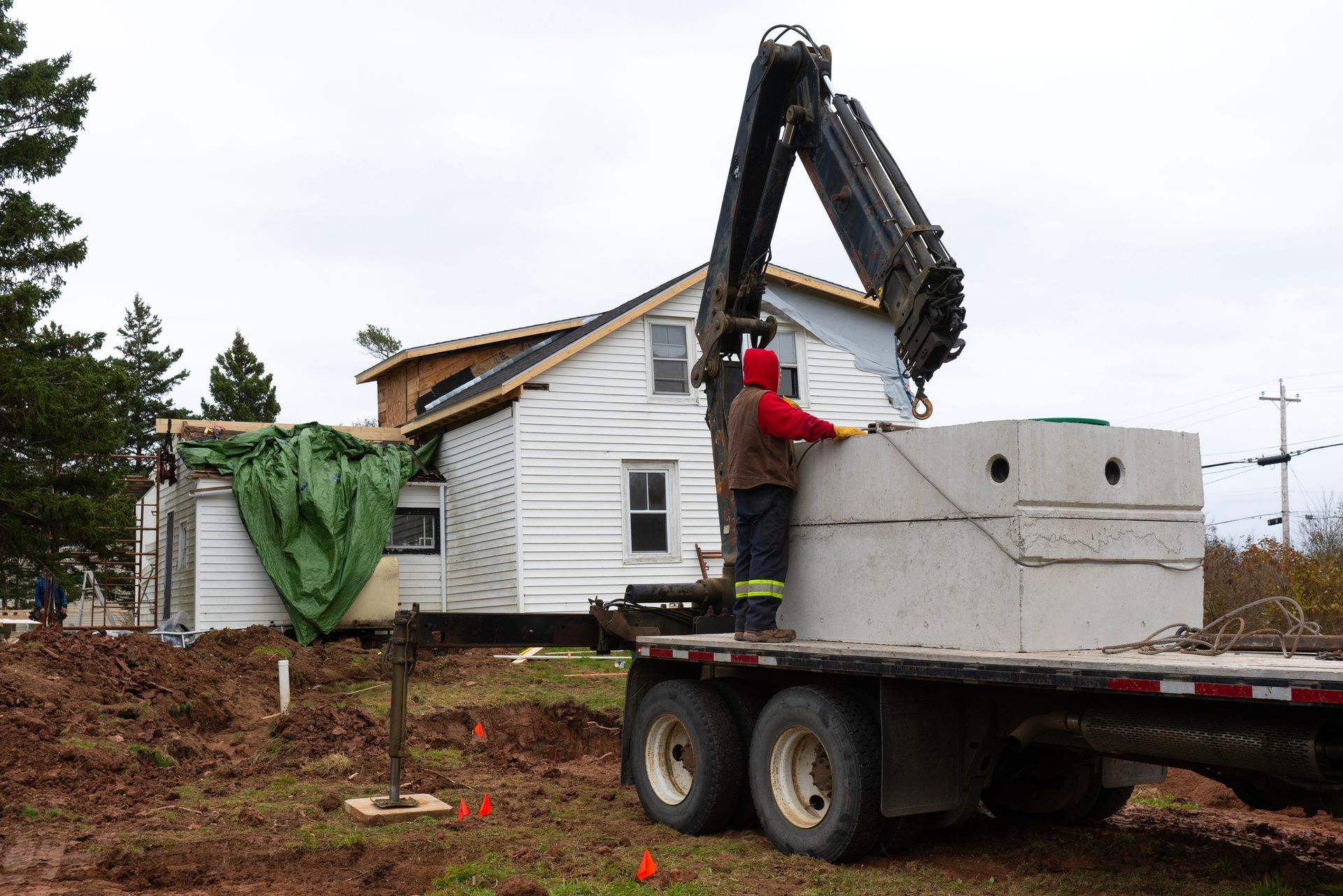 Crane lifts concrete septic tank from a truck near a white house undergoing construction. A worker guides the tank.
