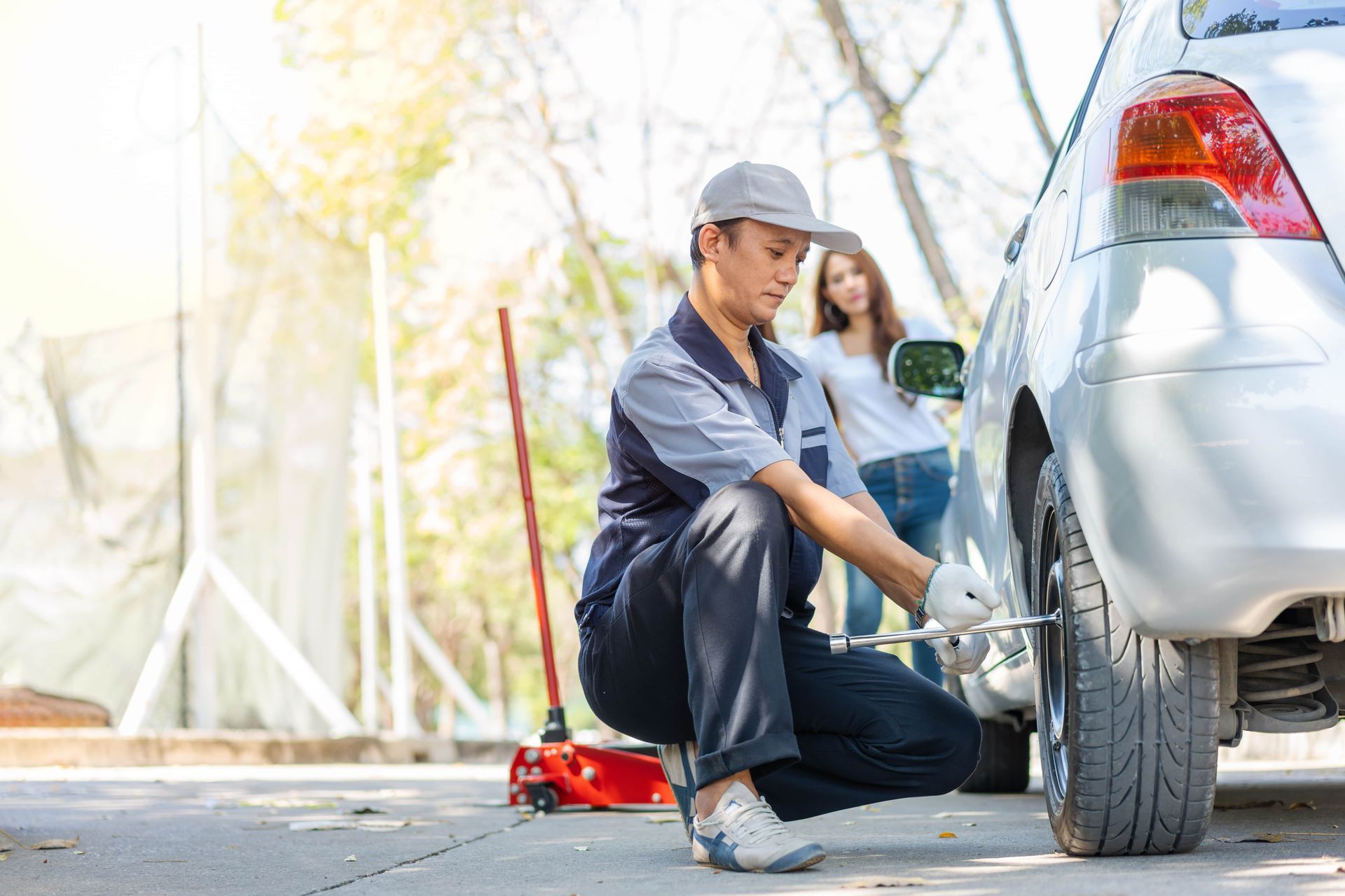 A mechanic in a grey uniform changing a car tire with a woman watching, on a sunny day.