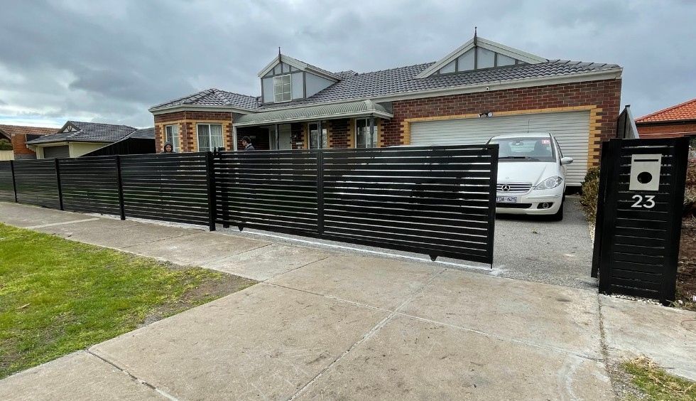 Black slatted fence and gate in front of a brick house with a white car in the driveway, cloudy day.