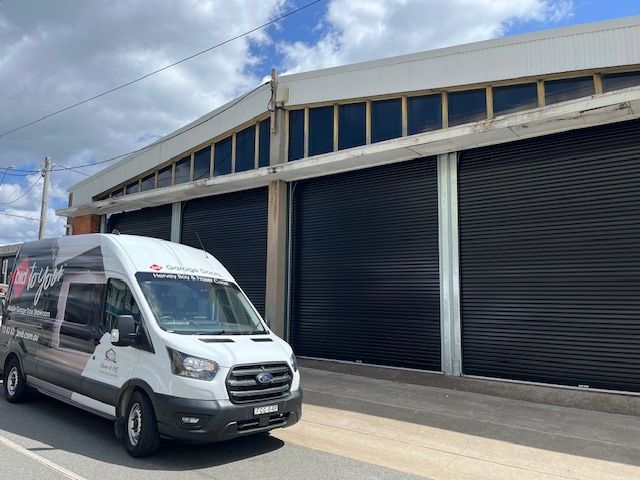 White van parked outside a building with black roller doors; the sky is blue with clouds.