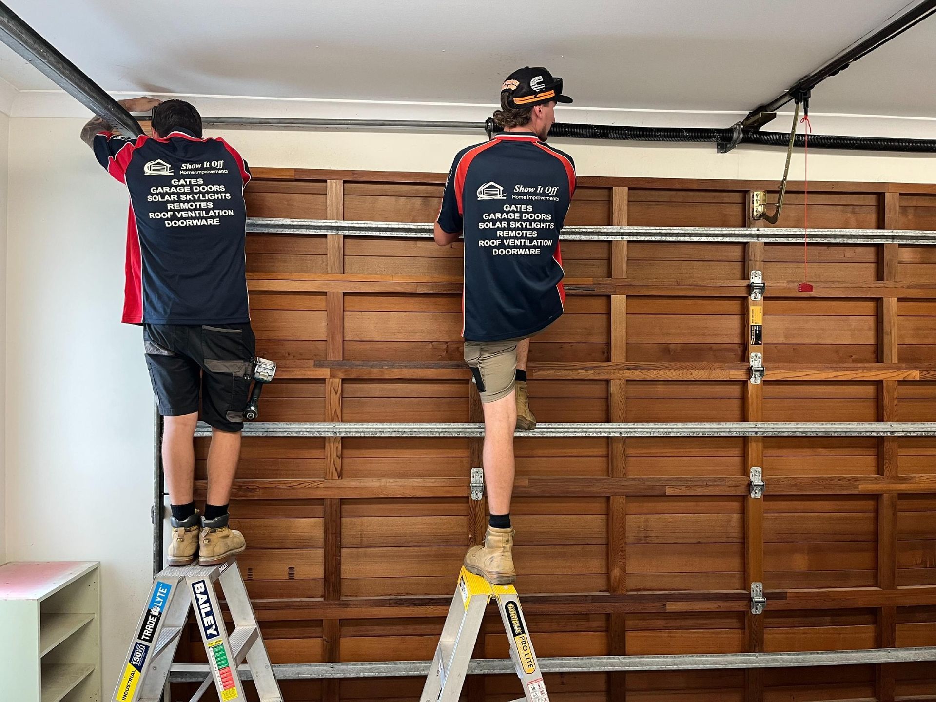 Two workers on ladders install garage door hardware inside a wooden garage.