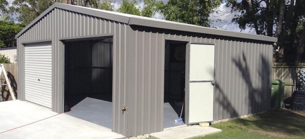 A grey metal shed with a garage door and a white door, set on a concrete slab, with grass and trees.
