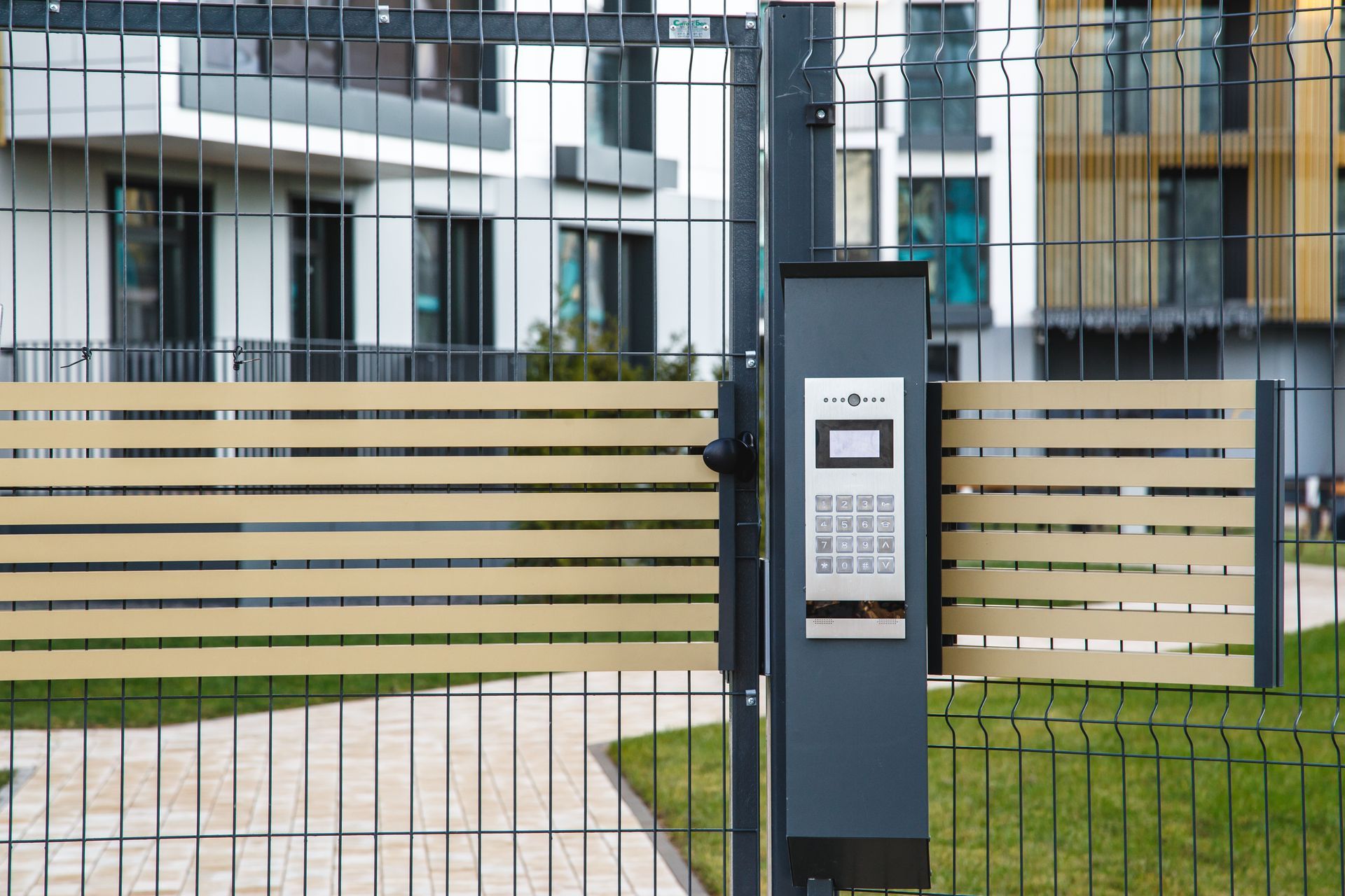 Black gate with keypad, beige horizontal slats, green grass, modern apartment buildings in the background.