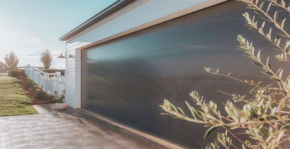 Dark gray garage door on a house with a white fence, and green grass.