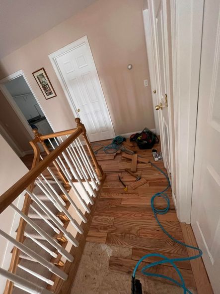 An indoor hallway with light-colored walls features a wooden staircase, closet doors, and partial hardwood floor installation.