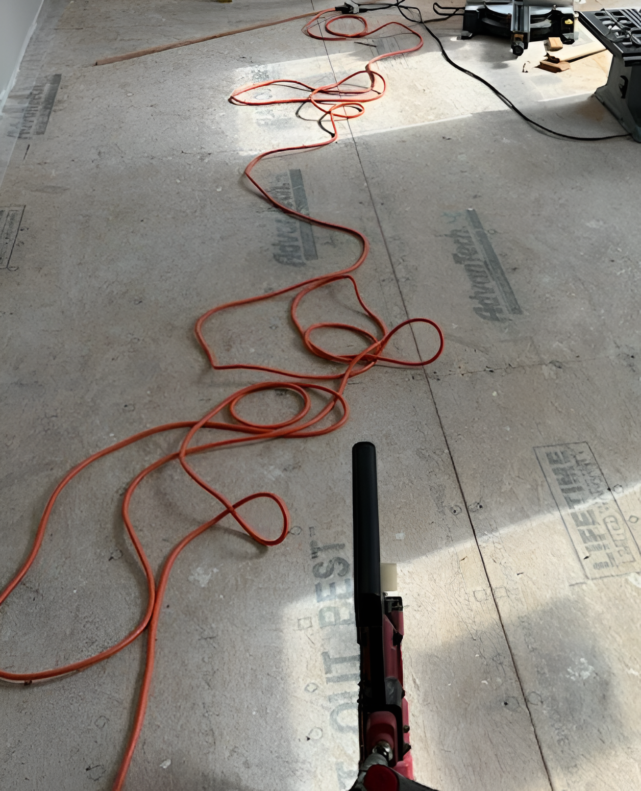 An orange extension cord snakes across a floor of subflooring panels toward a power tool in the foreground.