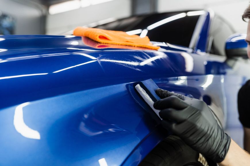Gloved hand polishing a glossy blue car body with an orange cloth at an auto shop