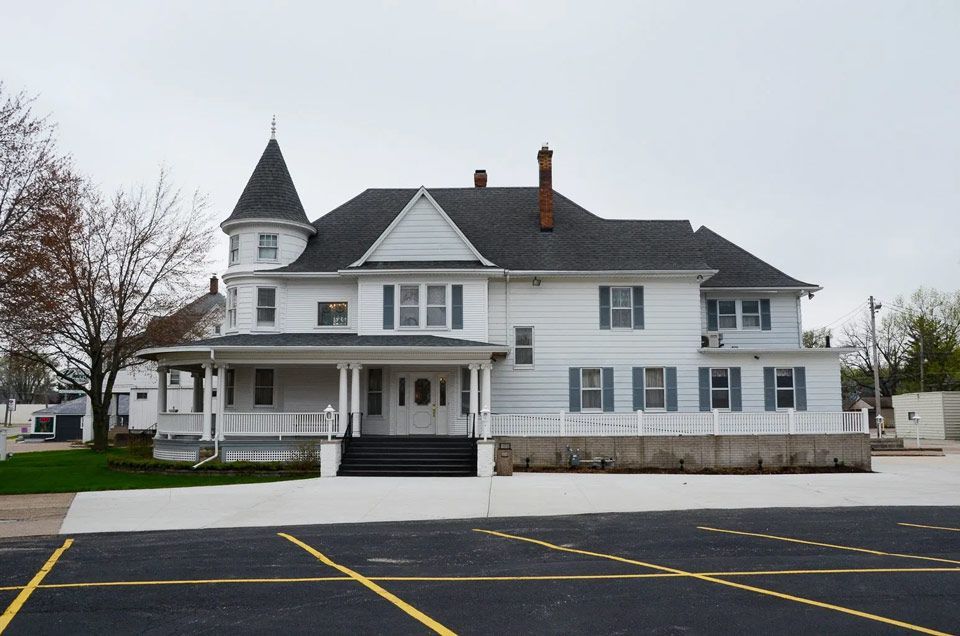 A large white house with a black roof sits in a parking lot