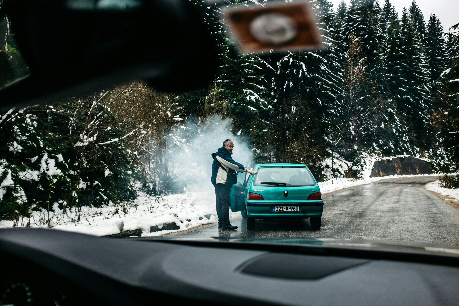 Een man staat naast een blauwgroene auto op een besneeuwde weg met rook. Bos op de achtergrond.