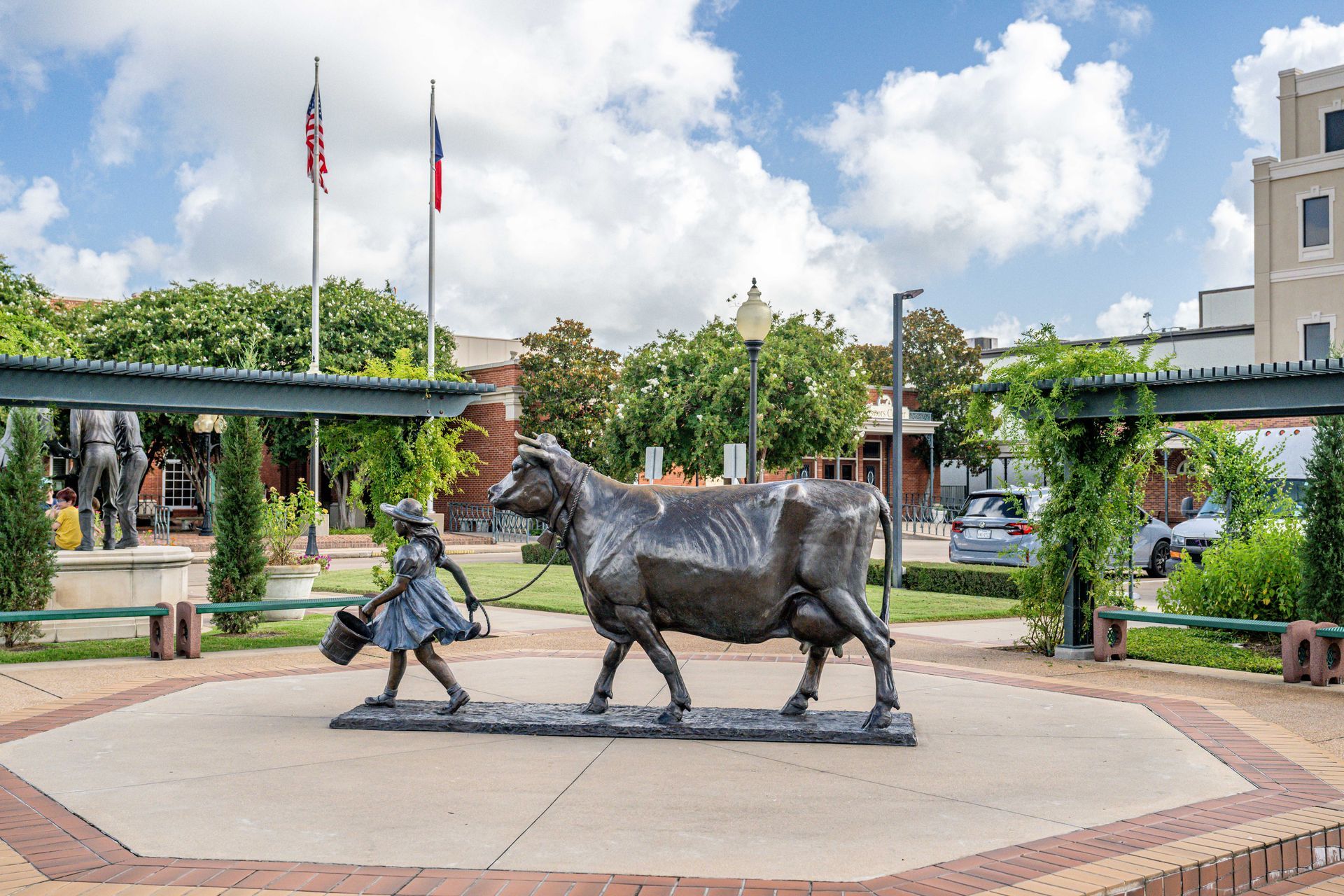 Statue of a girl leading a cow in a town square with flags and buildings.