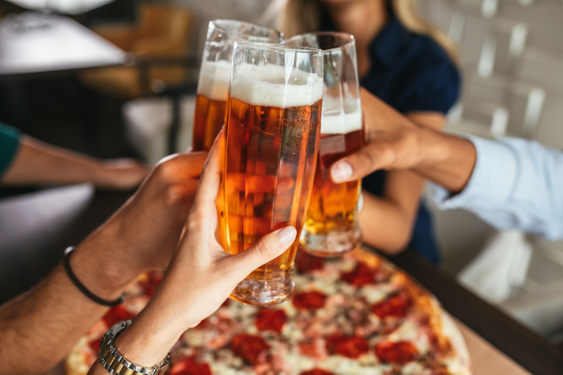 Hands holding beer glasses clinking together above a pizza on a wooden surface.
