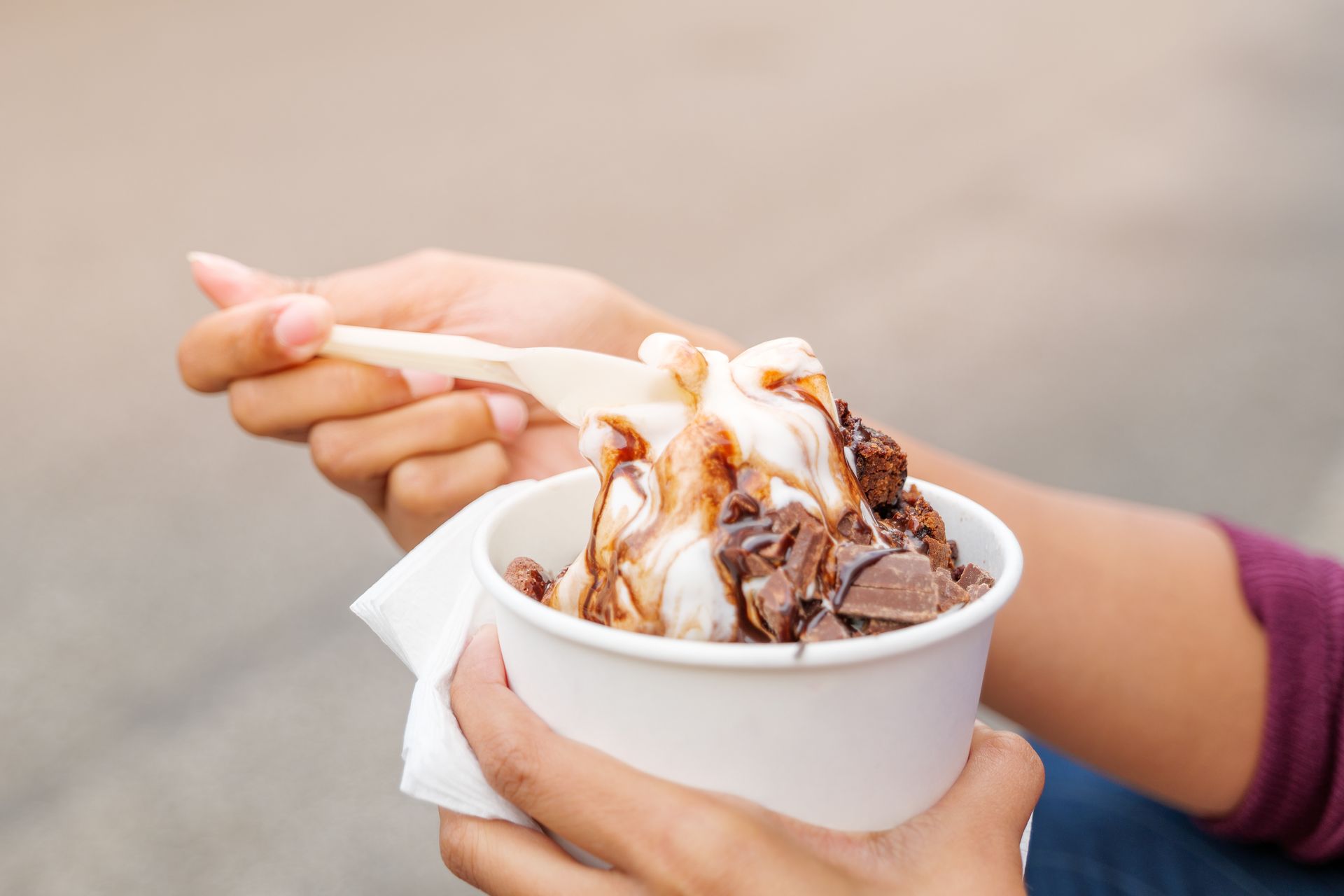 Person holding a cup of soft-serve ice cream with chocolate sauce and pieces, using a wooden spoon.
