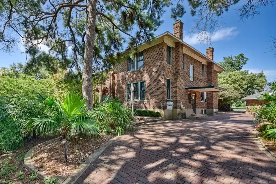 Brick house with a long driveway under the shade of trees and greenery.