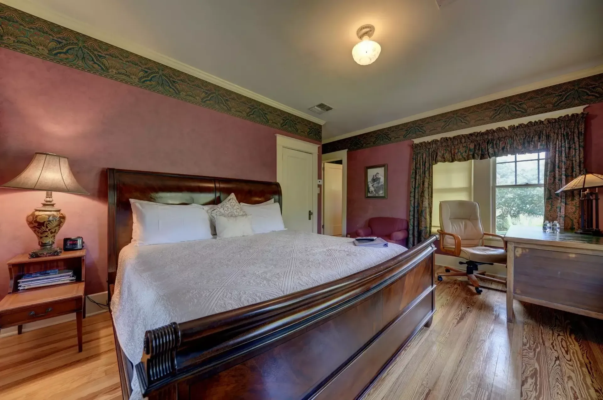 Bedroom with a large bed, nightstand, desk, and window. Walls are purple with decorative trim.