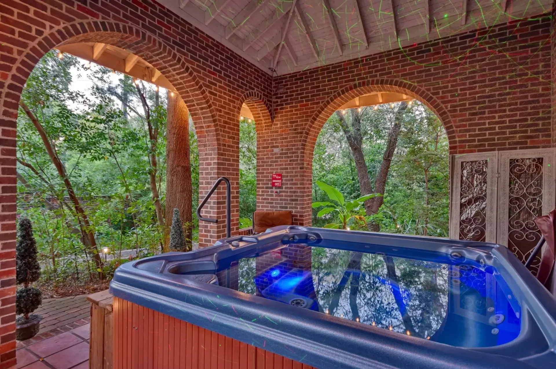 Hot tub under brick archway with views of lush green trees. Blue water reflects overhead lights.