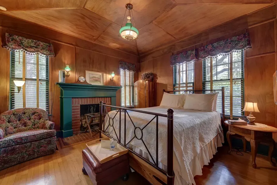 Bedroom with wooden walls, fireplace, bed, armchair, windows, and antique furnishings.