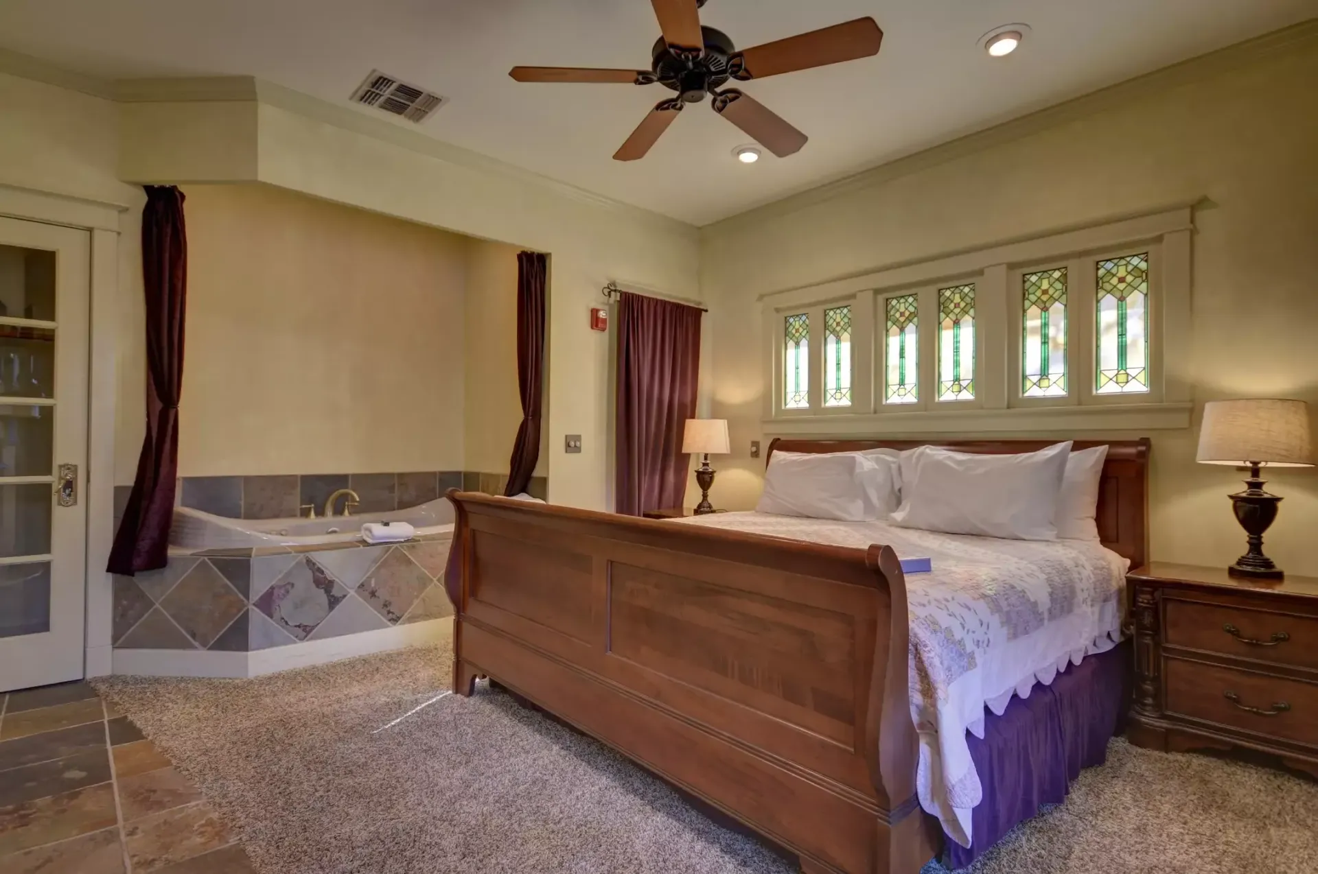 Bedroom with bed, jacuzzi, stained glass window, neutral colors, and ceiling fan.