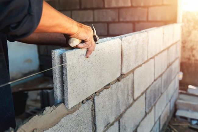 A worker lays a light-colored concrete block on a wall while using a string line as a guide.