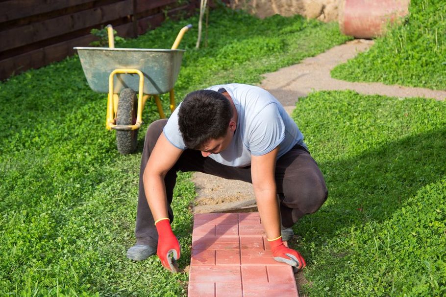 A person wearing red gloves crouches in a grassy yard, carefully placing red paving stones in a row near a wheelbarrow.
