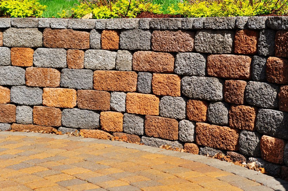 Curved stone retaining wall with a mix of tan and grey blocks, set behind a paved patio with greenery at the top.