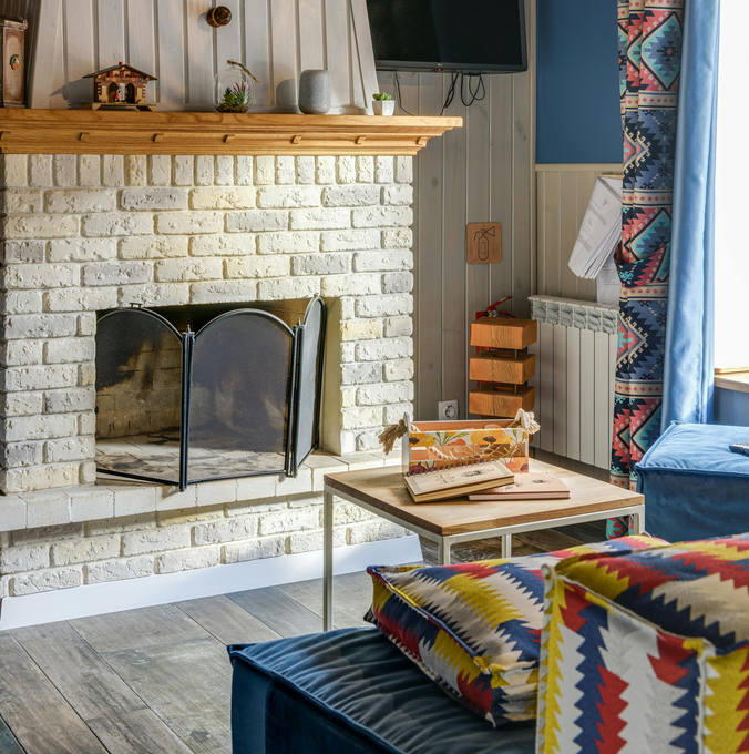 A brick fireplace with a metal screen and wooden mantel, next to a small table with books and patterned blue furniture.