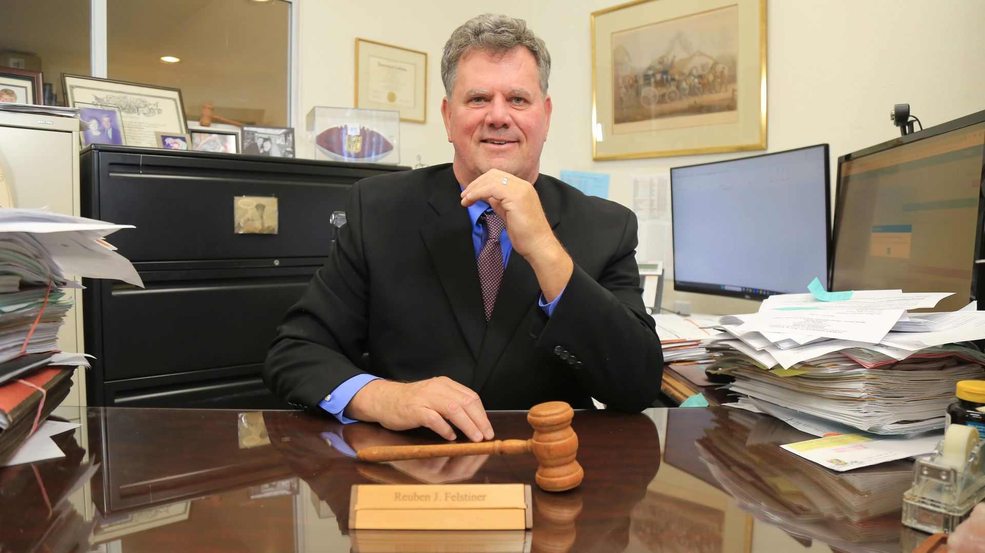 Man in suit seated at desk, holding gavel. Office setting, smiling, papers visible. Man in suit seated at desk, holding gavel. Office setting, smiling, papers visible.
