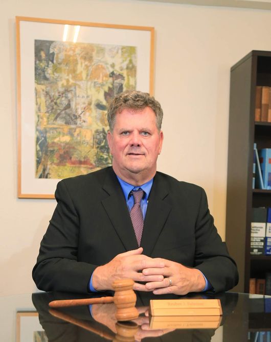Man in a suit behind a desk, hands clasped, next to a gavel and nameplate; painting on the wall. Man in a suit behind a desk, hands clasped, next to a gavel and nameplate; painting on the wall.