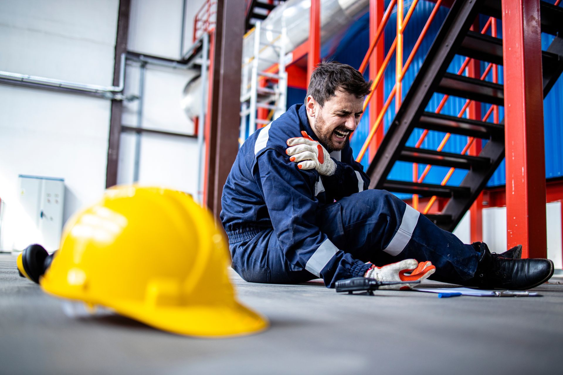 Injured worker sitting on the floor holding a shoulder, with tools and a helmet nearby. Injured worker sitting on the floor holding a shoulder, with tools and a helmet nearby.