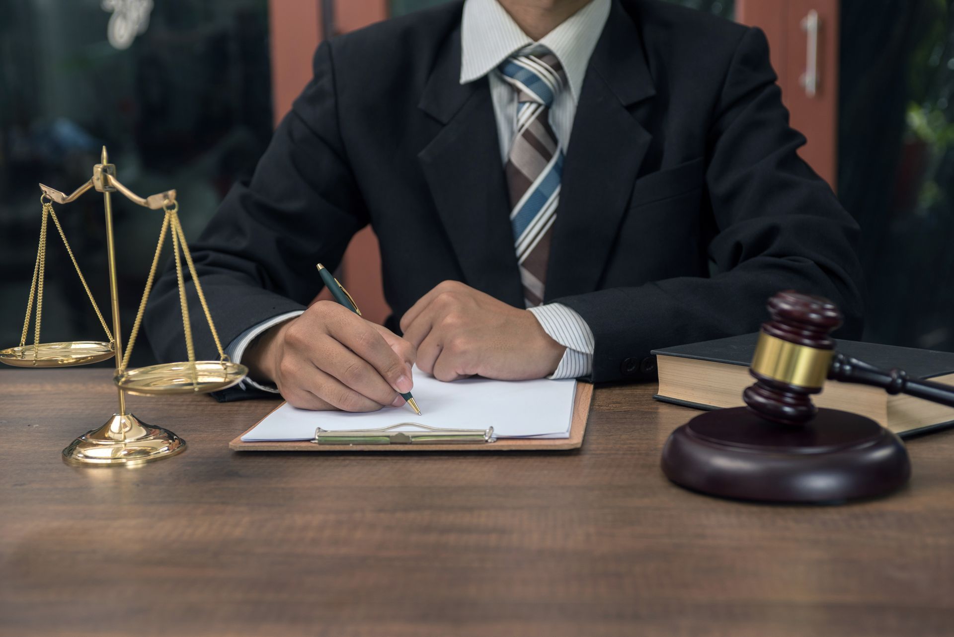 A person in a suit writes at a desk with a gavel, law books, and scales of justice. A person in a suit writes at a desk with a gavel, law books, and scales of justice.