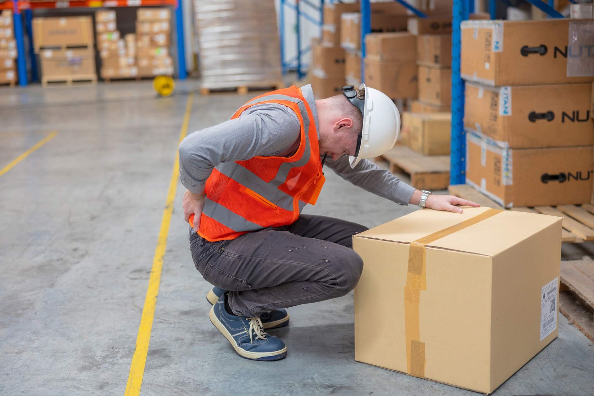Warehouse worker in safety vest holding back, squatting by cardboard box. Warehouse worker in safety vest holding back, squatting by cardboard box.