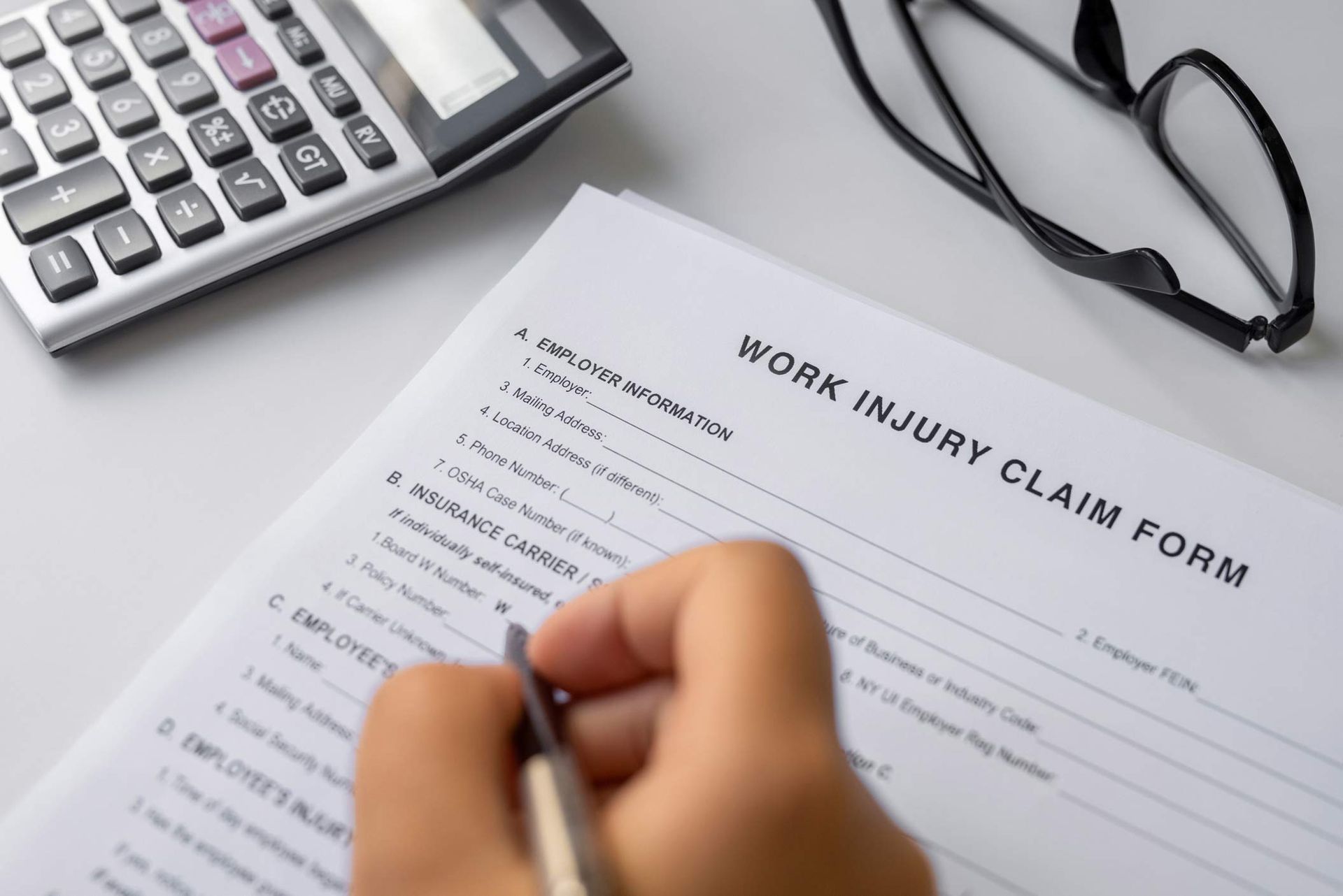 Person filling out a work injury claim form, with a calculator and glasses on a table. Person filling out a work injury claim form, with a calculator and glasses on a table.