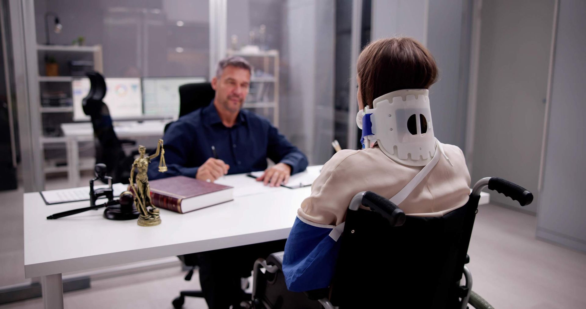 A person in a wheelchair and neck brace meets with a lawyer in an office. A person in a wheelchair and neck brace meets with a lawyer in an office.
