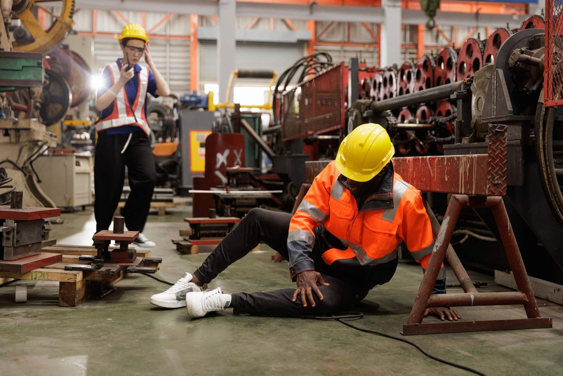 Worker fallen on the factory floor; another worker approaches with a flashlight, wearing safety gear. Worker fallen on the factory floor; another worker approaches with a flashlight, wearing safety gear.