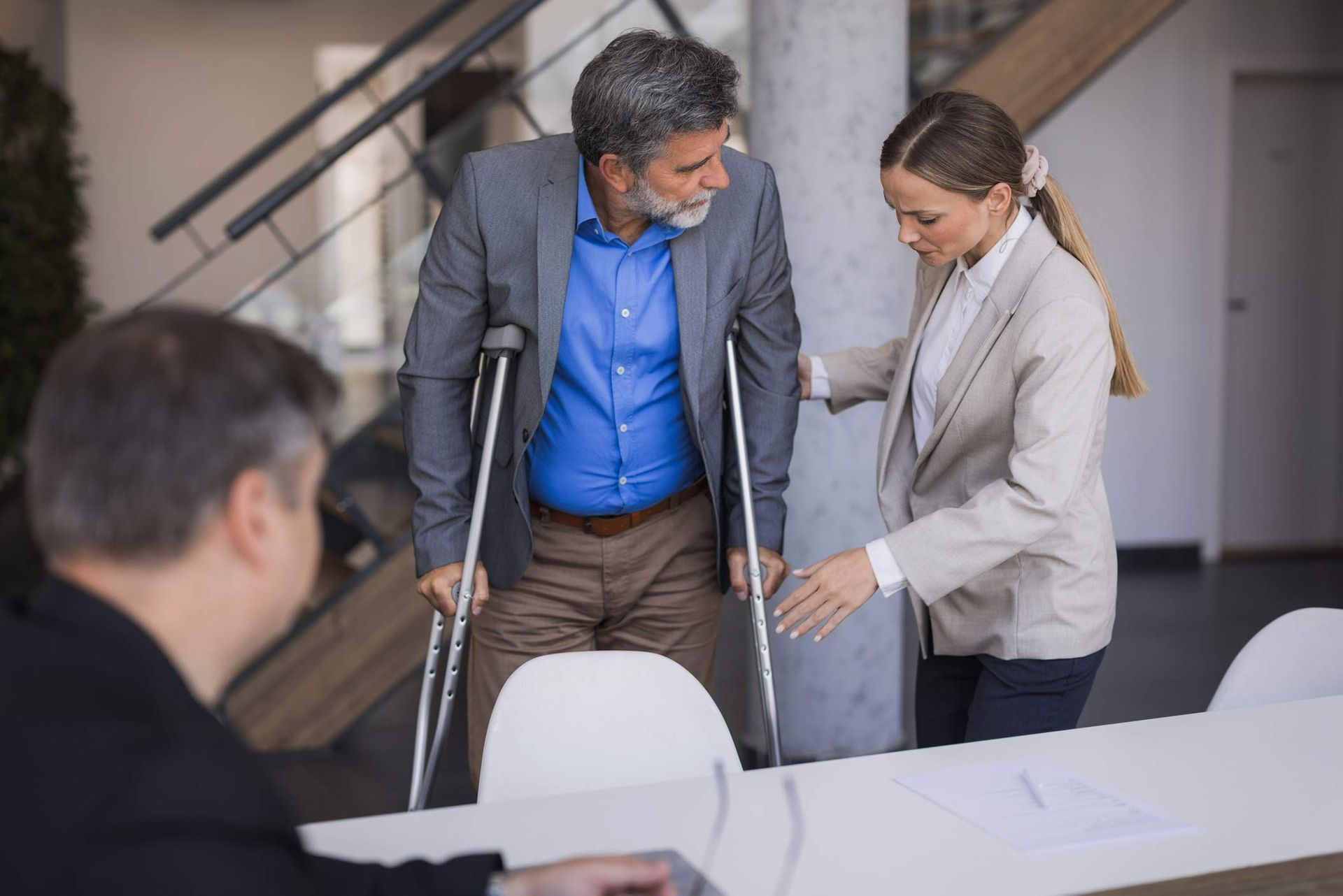 Man with crutches assisted by woman in office setting; another person sits at a table. Man with crutches assisted by woman in office setting; another person sits at a table.