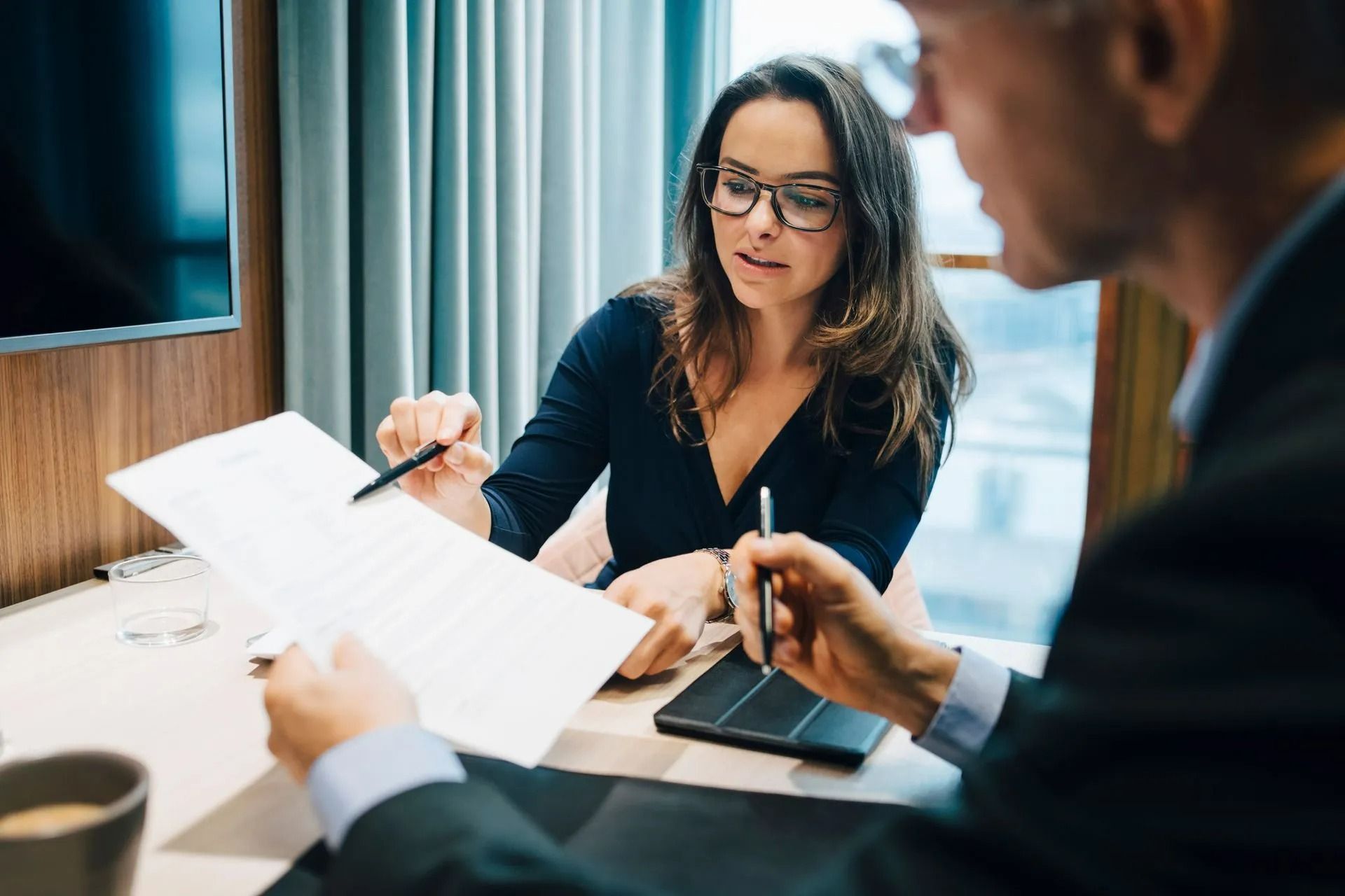 Woman in glasses points at document, reviewing it with a man in a business setting.