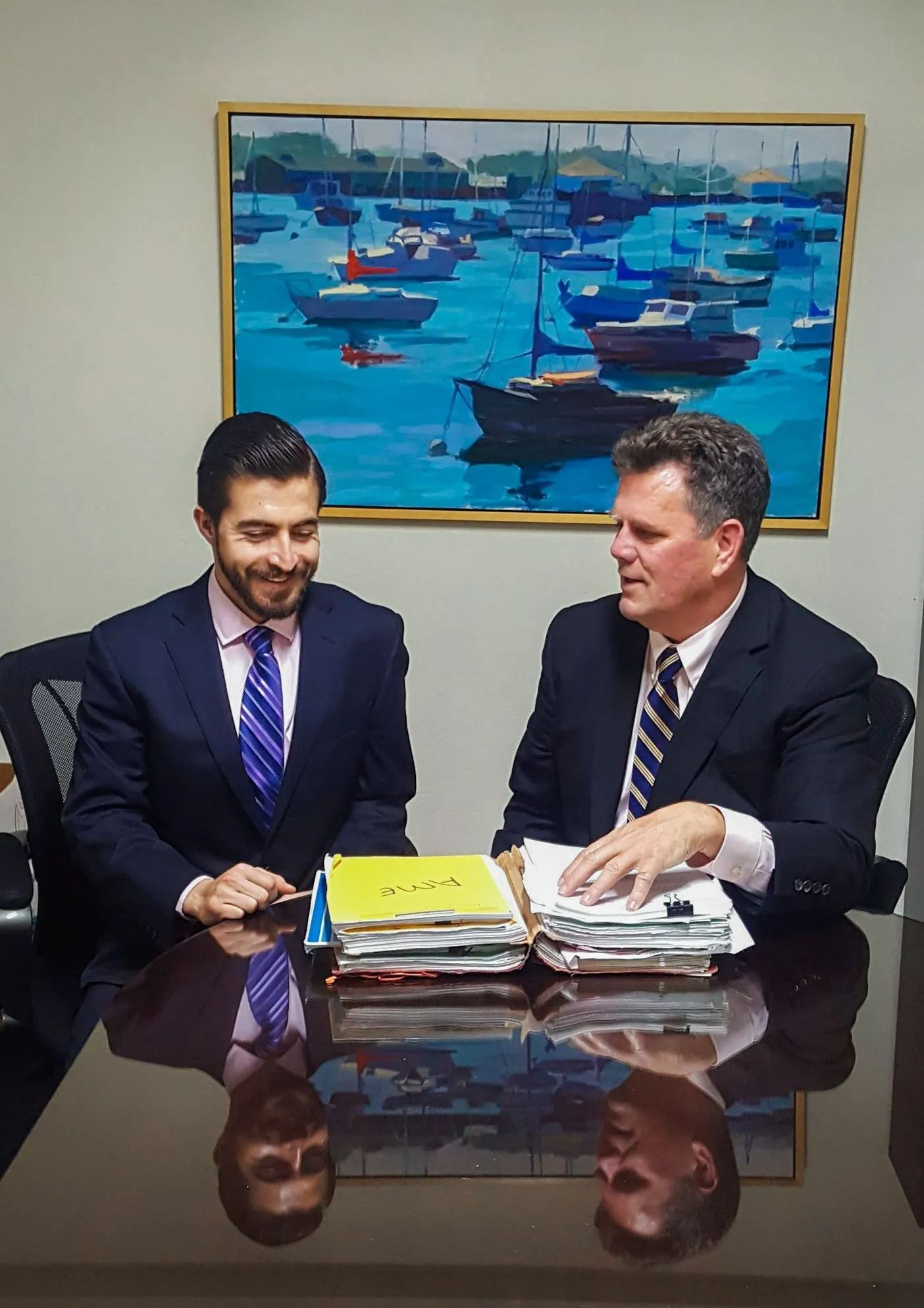 Two men in suits at a desk with papers, smiling, discussing. Blue harbor painting on wall.