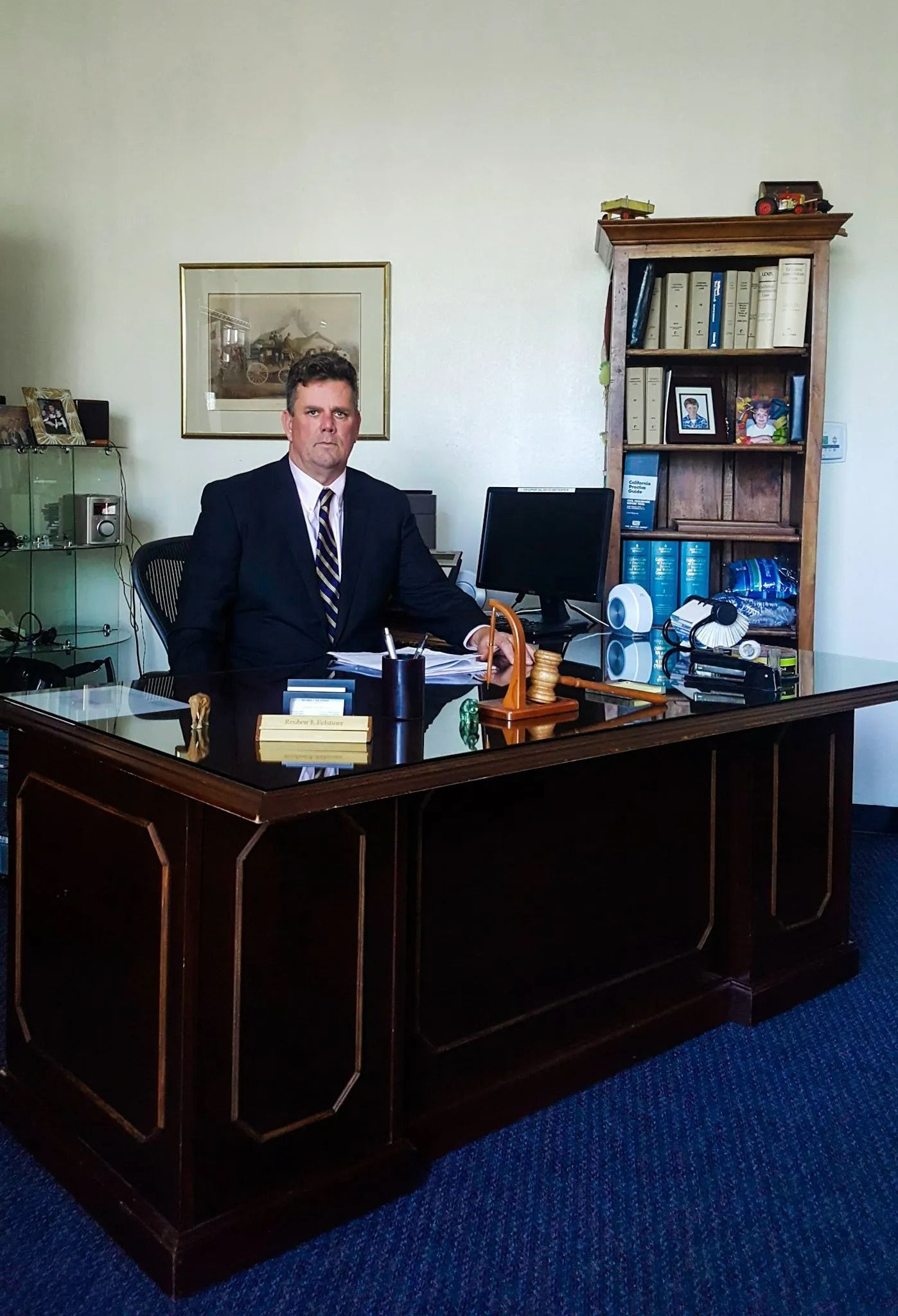 Man in a suit sits at a large desk in an office with a bookcase and artwork. Man in a suit sits at a large desk in an office with a bookcase and artwork.