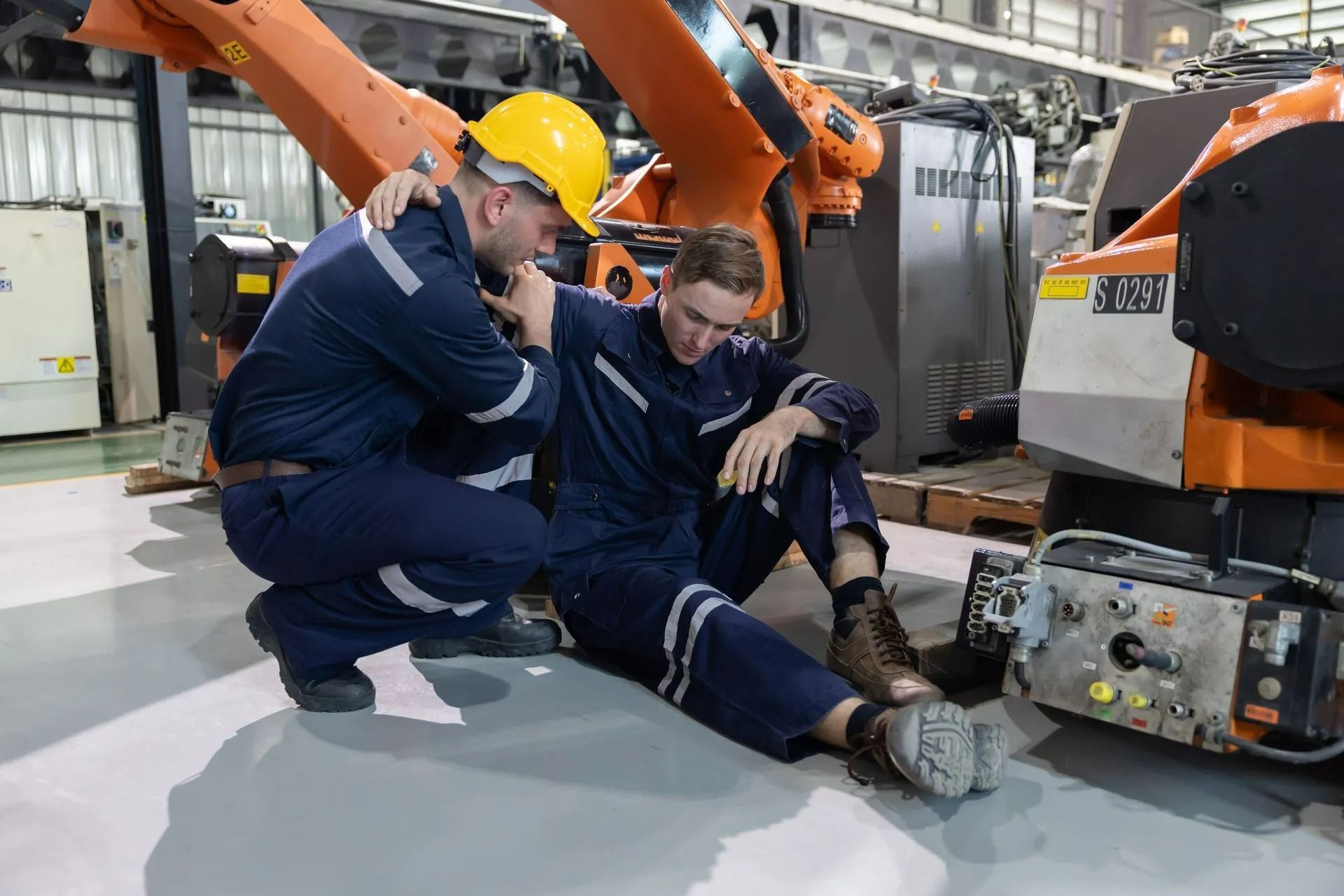 Worker helps injured colleague on factory floor near orange robot arm.
