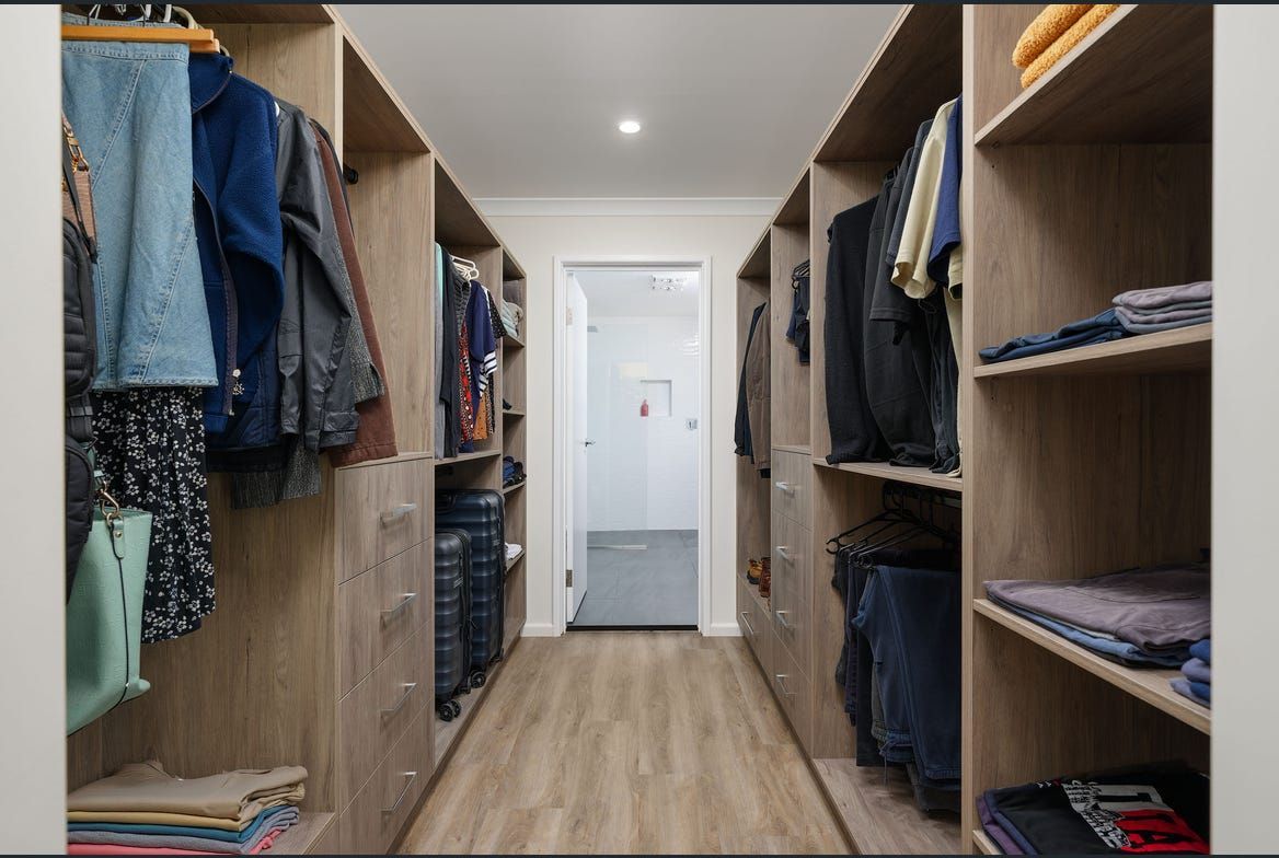 Walk-in closet with light wood shelving, clothes hanging and folded, and a door at the end — Morrison Cabinetry in Sunshine Coast, QLD