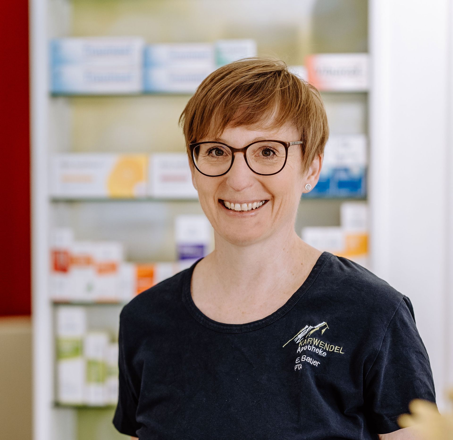 Eine Frau mit Brille und schwarzem Hemd lächelt in einer Apotheke.