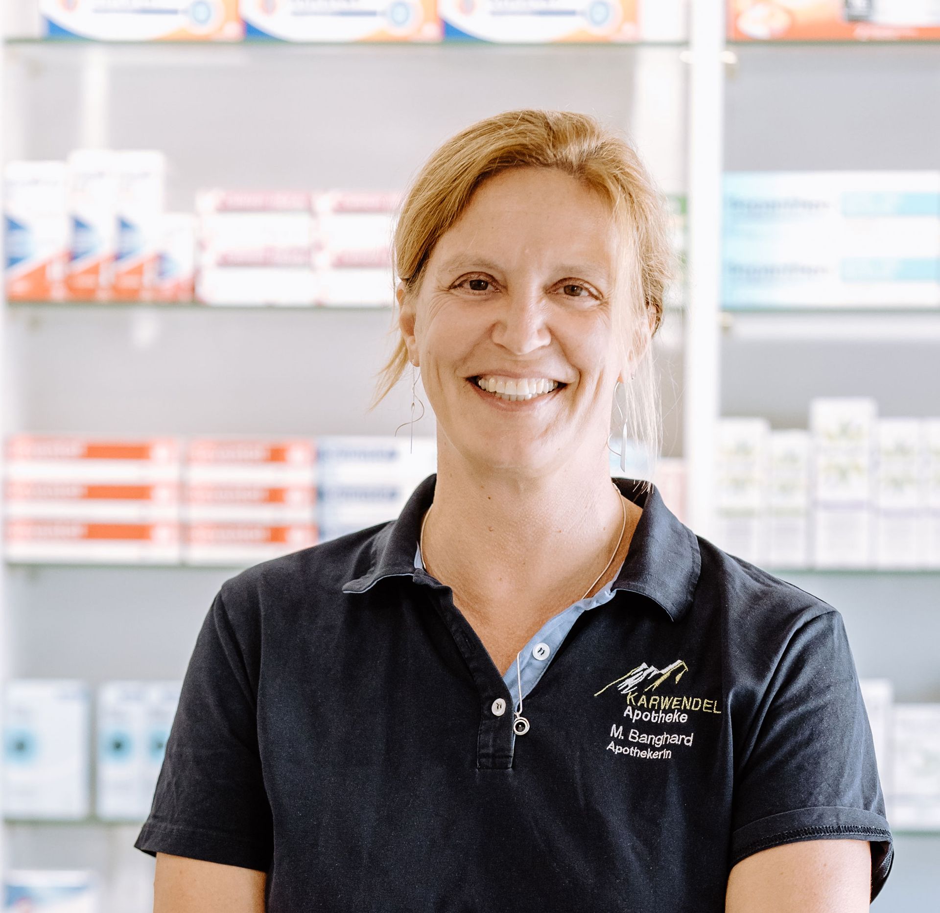 Eine Frau in einem schwarzen Hemd lächelt vor einem Apothekenregal.