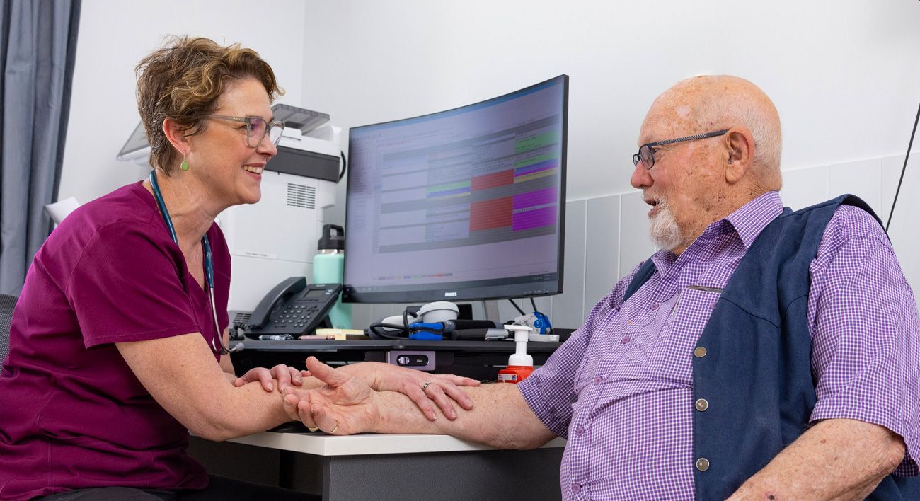 An elderly man is sitting at a desk talking to a nurse.